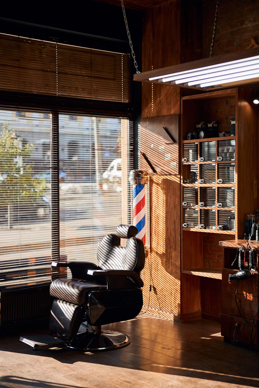 Silla de barbero de cuero junto a una ventana con persiana de madera, con el poste de barbería y los estantes de herramientas al fondo