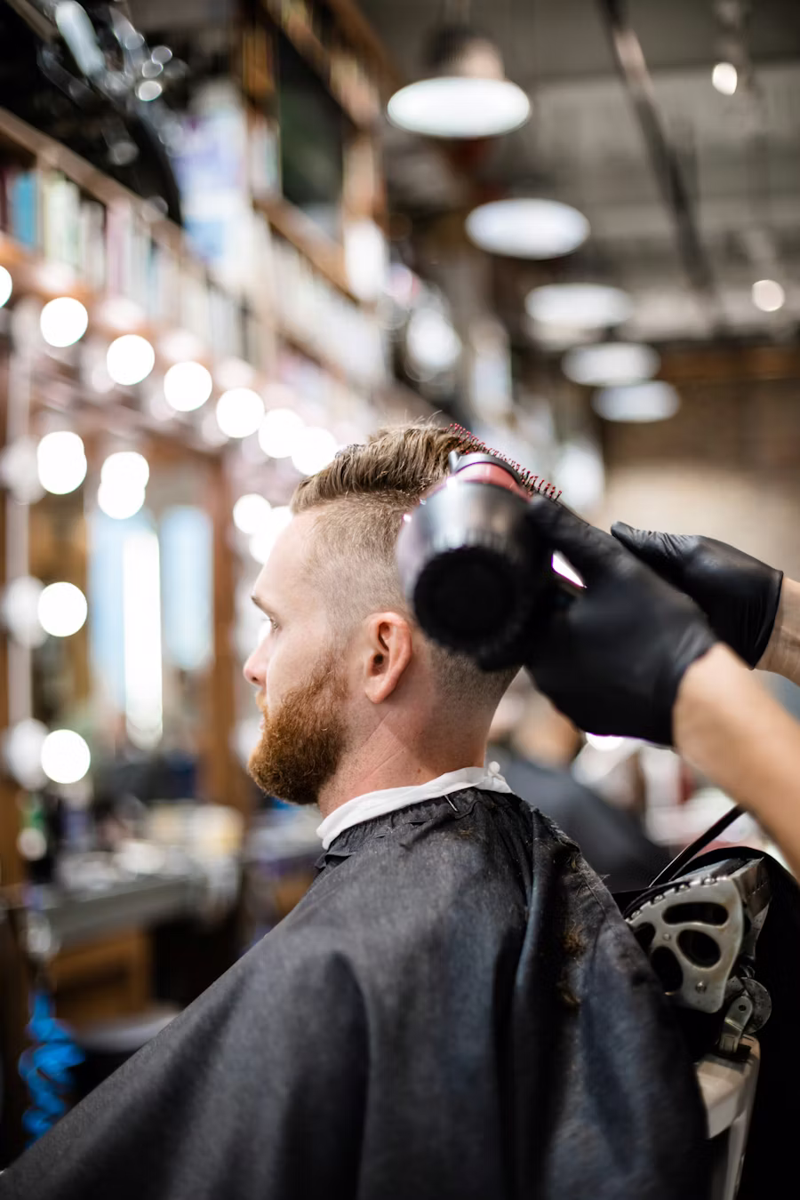 Barbero secando el pelo de un cliente de espaldas, frente al espejo con bombillas del local